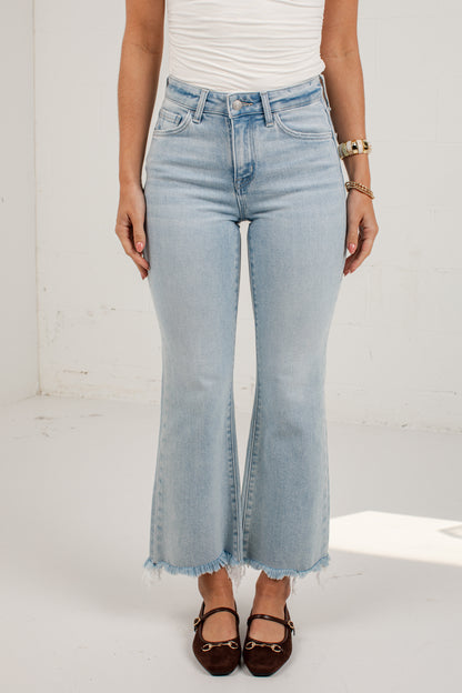 A model wearing light-wash blue flared jeans with a raw frayed hem, paired with a white top and dark brown flat shoes, standing against a white wall.