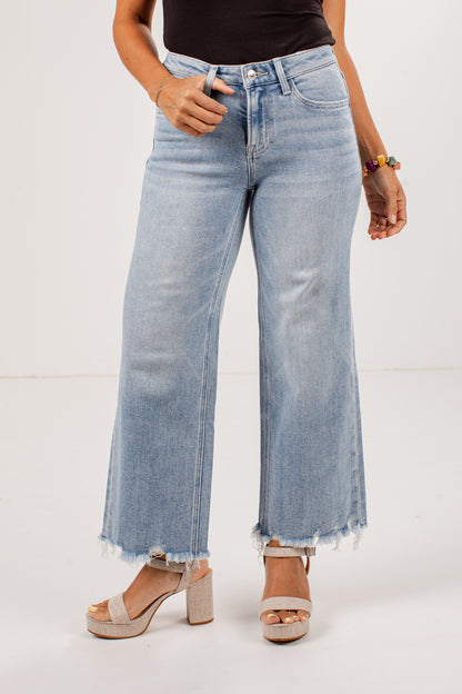 A model wearing light-wash blue wide-leg cropped jeans with a raw frayed hem, paired with beige platform sandals and a black top, shown against a plain white studio background.
