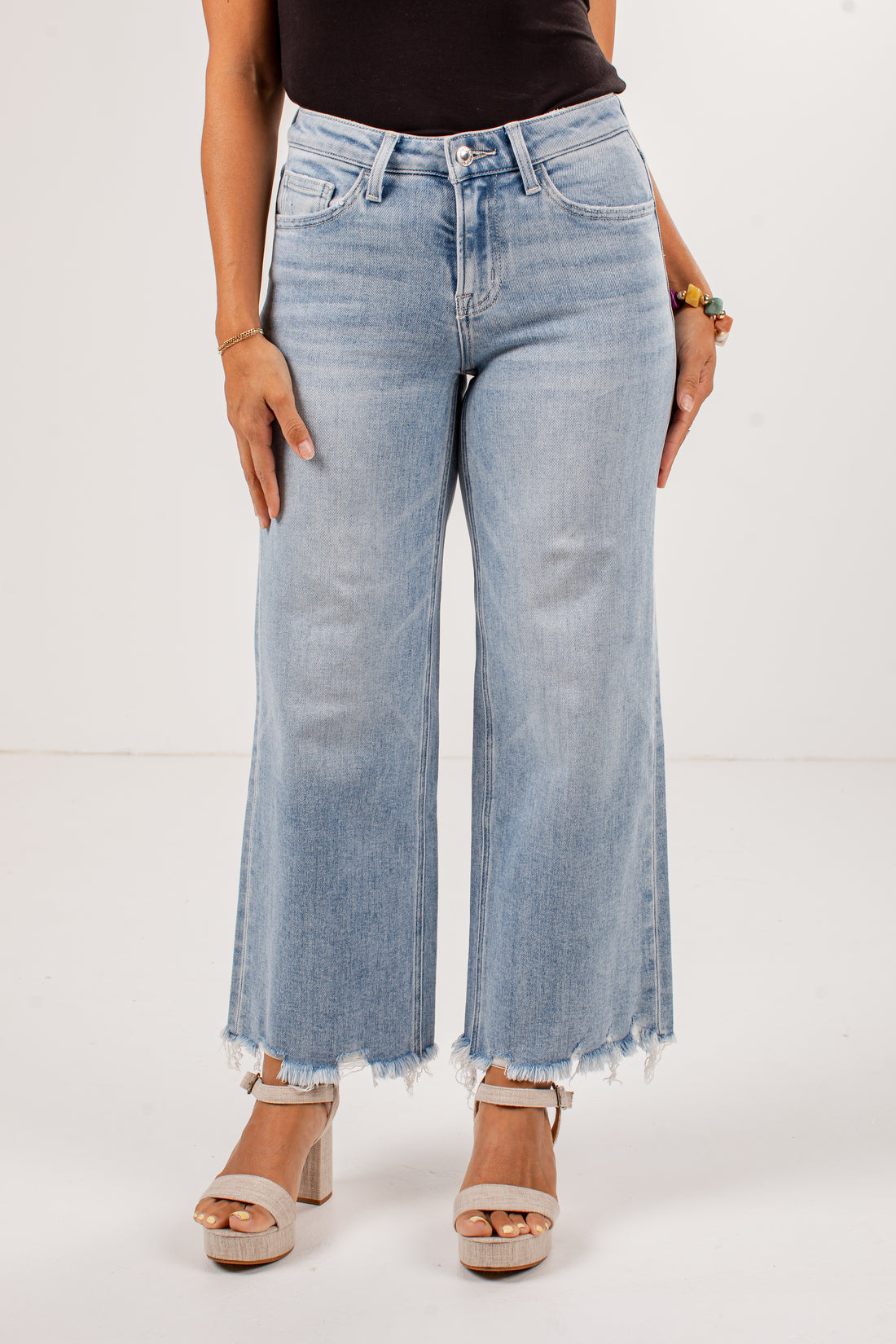 A model wearing a pair of light-wash blue wide-leg cropped jeans with a raw frayed hem, paired with beige platform sandals against a plain white studio background.