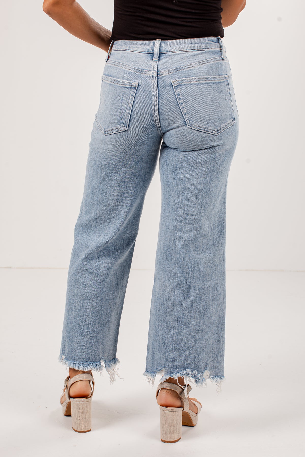 A person shown from the back wearing light-wash blue wide-leg cropped jeans with a frayed hem, paired with beige block-heel sandals against a plain light background.
