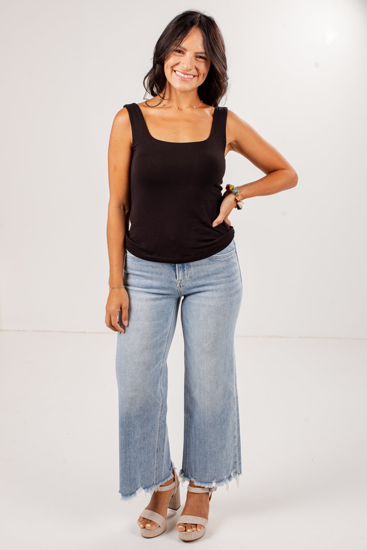 A woman modeling a black sleeveless tank top and light-wash wide-leg jeans with frayed hems, paired with beige heeled sandals, against a plain white studio background.