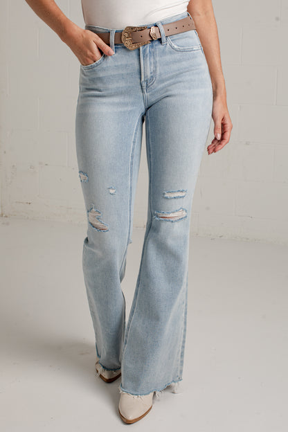 A person wearing light-wash blue distressed flare jeans with a brown belt, paired with a white top and beige pointed-toe heels, standing against a light gray wall.
