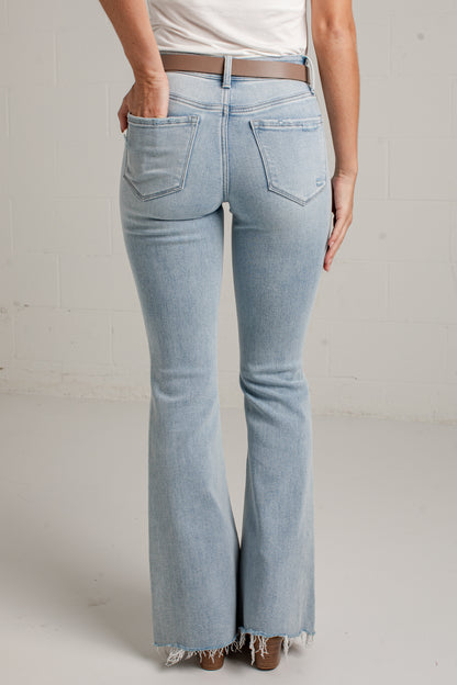 Rear view of a person wearing light-wash blue flare jeans with a frayed hem, paired with a white top and brown belt, against a light gray wall.