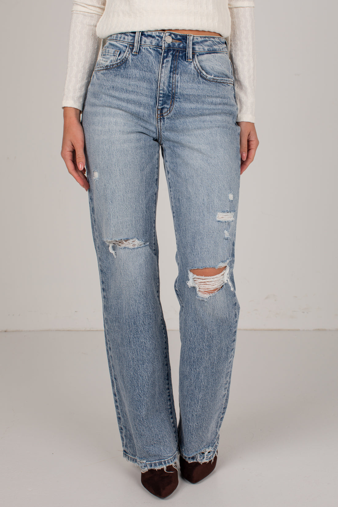 Light-wash blue distressed jeans with ripped details on the thighs and knees, shown on a model wearing a white ribbed long-sleeve top and dark brown pointed-toe heels against a plain light gray studio background.