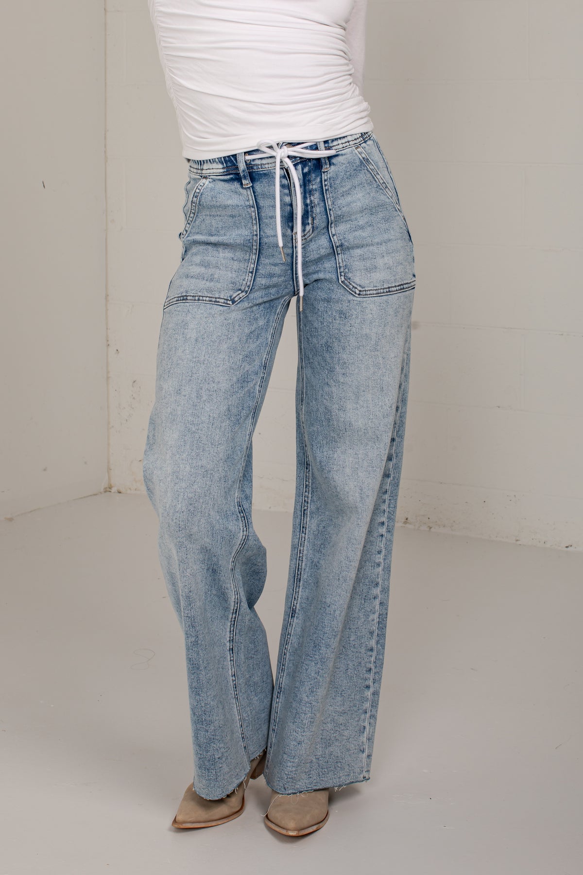 A person wearing light-wash wide-leg denim jeans with a drawstring waist, paired with a white top and tan pointed shoes, standing in a plain indoor studio corner.