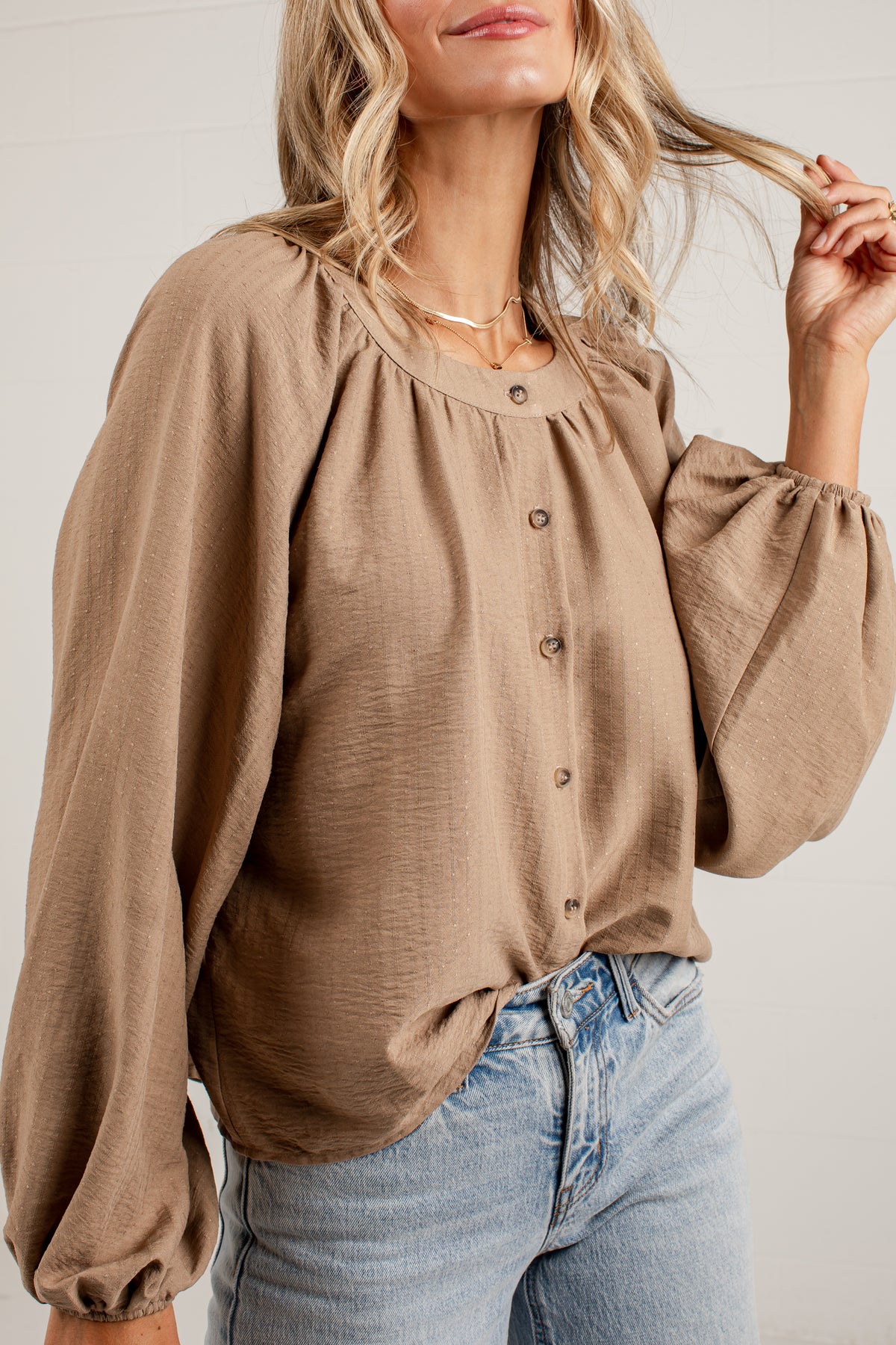 A woman wearing a taupe brown long-sleeve blouse with a gathered neckline and button-front detail, paired with light-wash blue jeans against a white paneled wall.