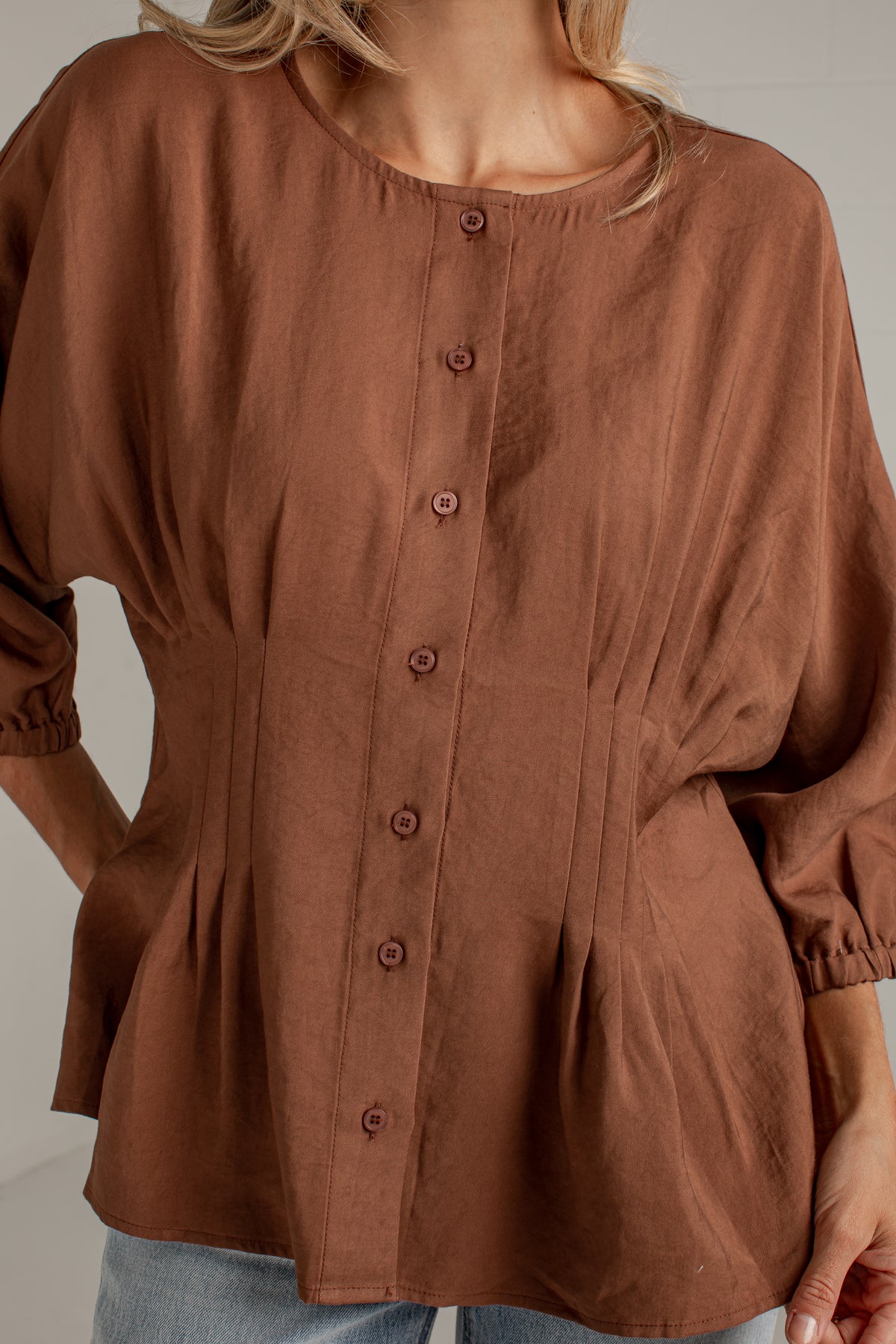A woman wearing a brown long-sleeve button-front blouse with a round neckline and gathered peplum hem, paired with light blue jeans.