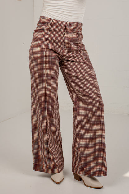 A person wearing high-waisted, wide-leg trousers with a brown and white houndstooth pattern, styled with a white top and white heeled mules.