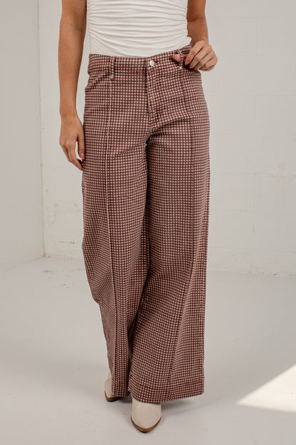 A person wearing high-waisted, wide-leg trousers in a brown and beige houndstooth pattern, paired with a white ribbed top and beige boots, standing against a white studio wall.