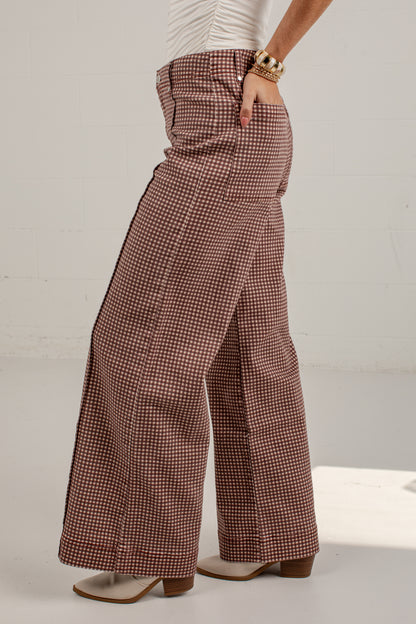 A person wearing high-waisted, wide-leg trousers in a small-scale brown and white houndstooth pattern, paired with a white top and beige block-heel sandals.