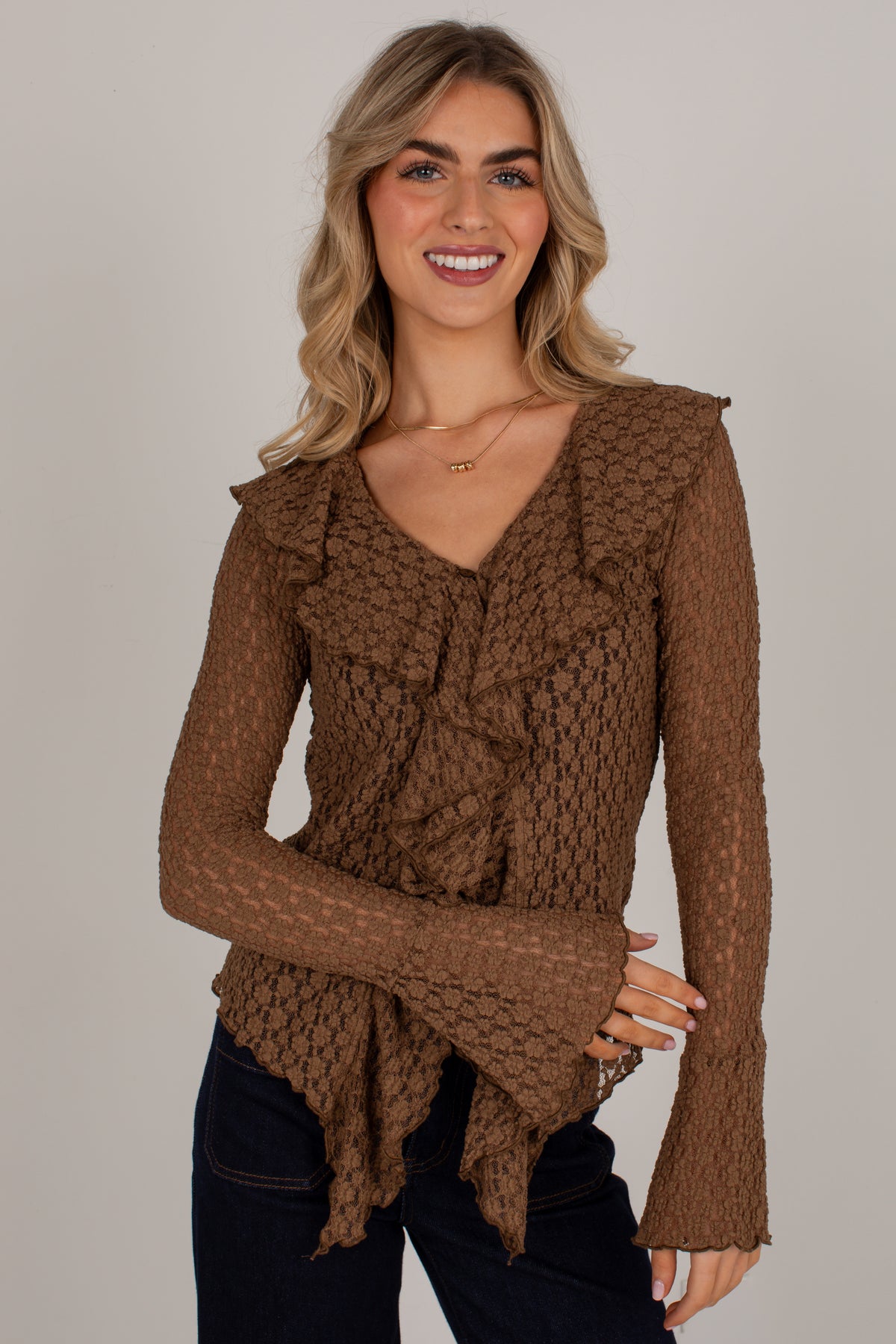 A woman wearing a brown textured knit long-sleeve top with ruffle details and a split neckline, paired with dark jeans, against a plain light gray background.