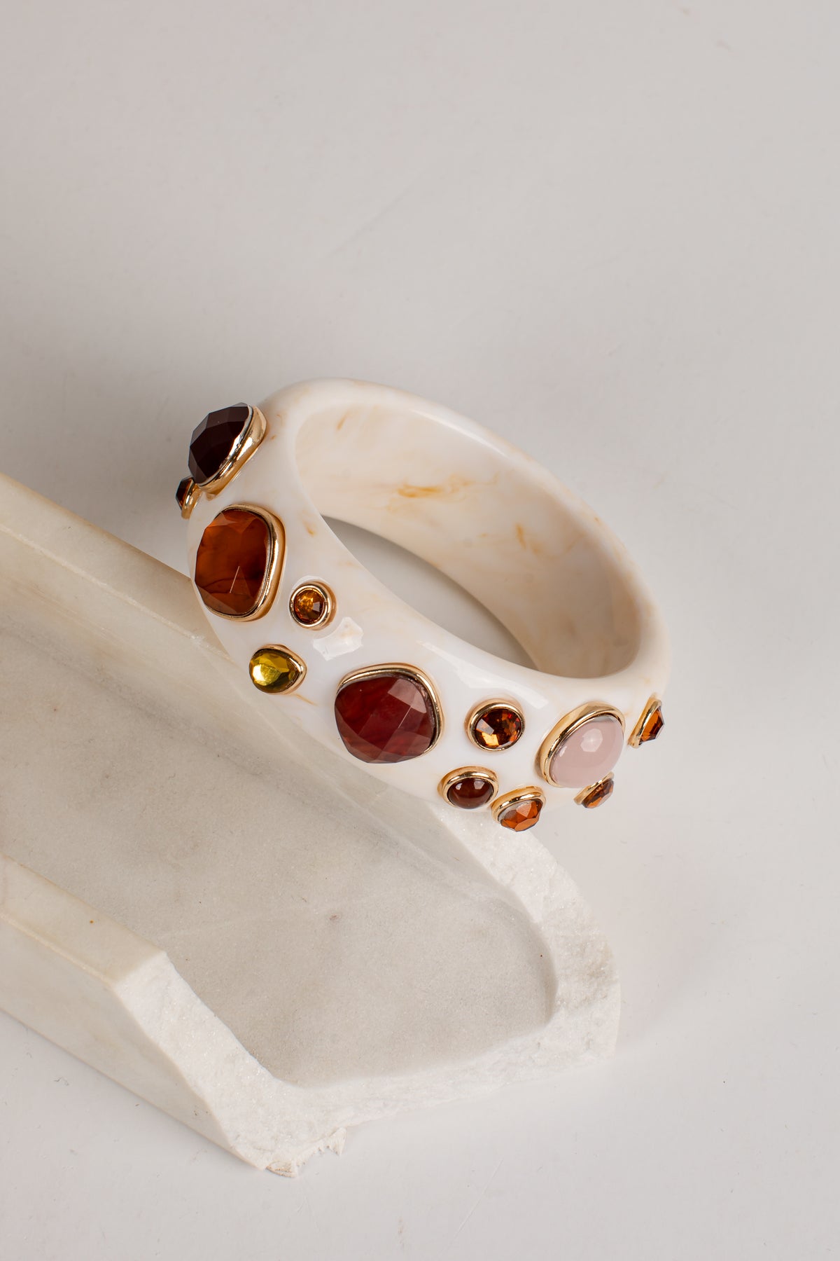 A cream-colored carved stone cuff bracelet decorated with multiple gold-set gemstones in various colors, displayed on a matching cream stone stand against a light gray background.