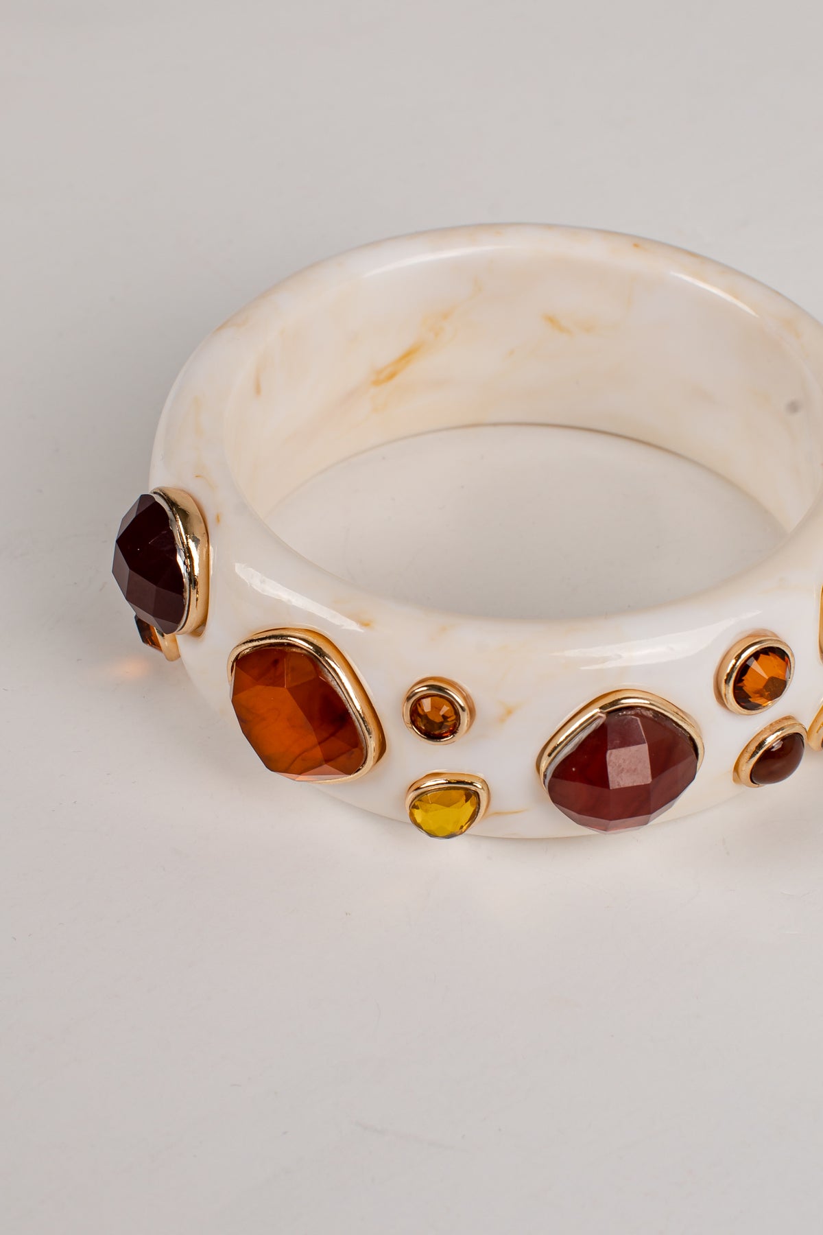 A white faux-marble bangle bracelet with gold-tone bezel settings holding faceted gemstones in shades of amber, burgundy, and yellow, shown on a light gray background.
