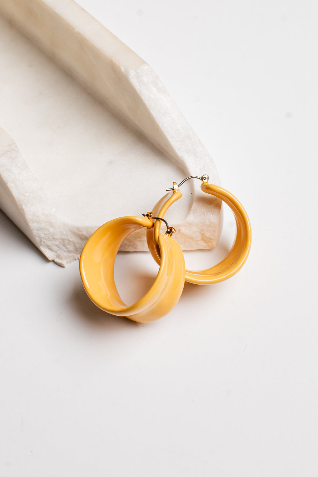 A pair of chunky, open hoop earrings in a glossy mustard yellow finish, resting on a textured white stone prop against a clean white background.