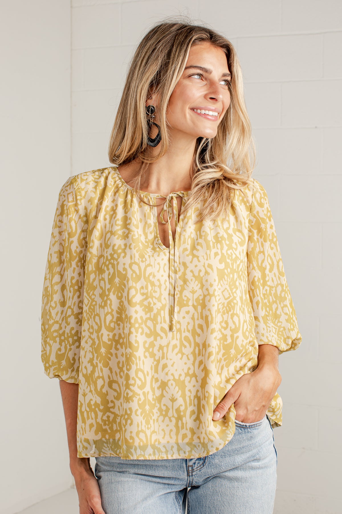 A woman wearing a yellow and cream patterned blouse with three-quarter sleeves and a tie neckline, paired with light-wash jeans, standing against a white brick wall.