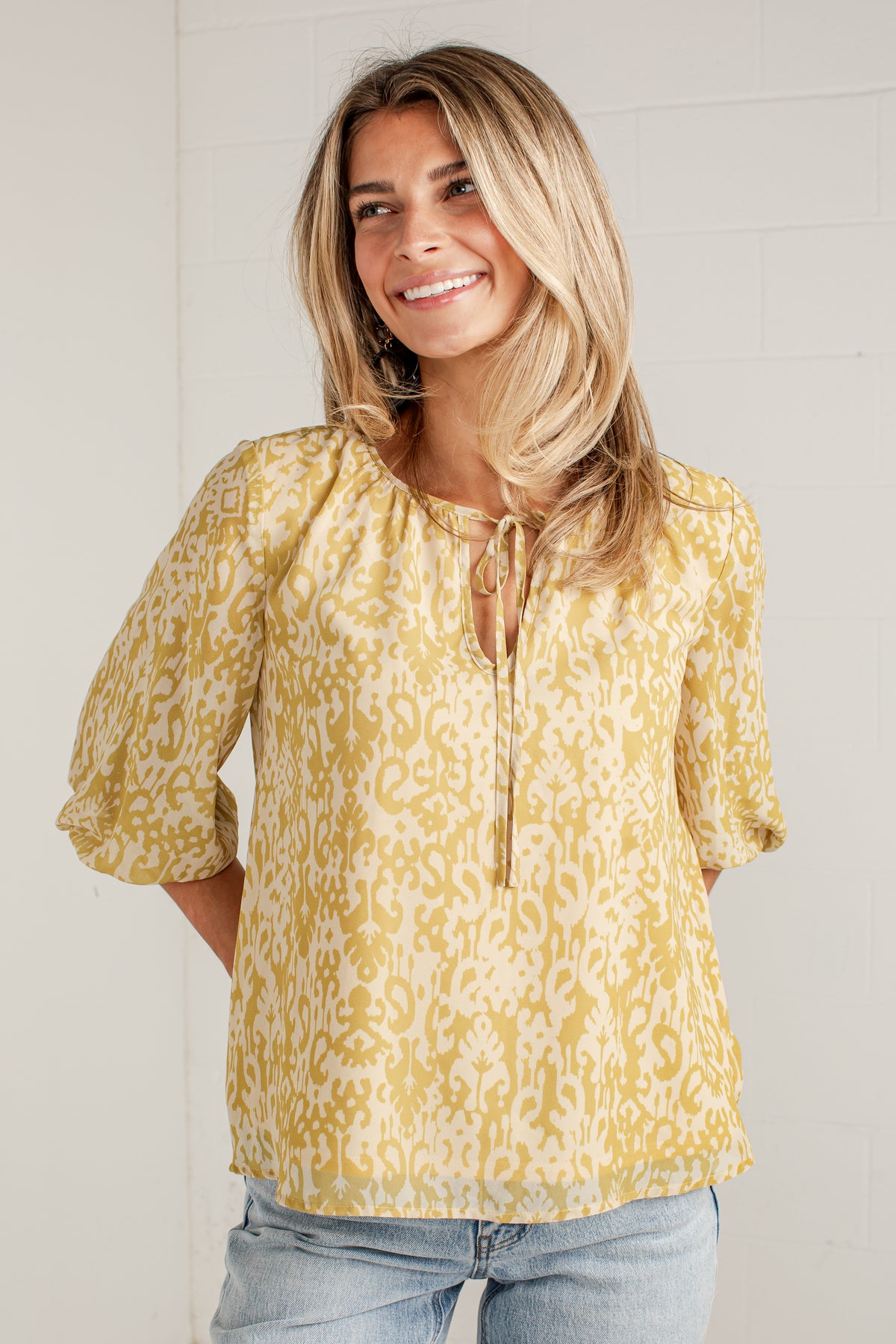 A woman wearing a yellow and cream patterned blouse with three-quarter sleeves and a tie neckline, paired with light blue jeans, standing against a white brick wall.