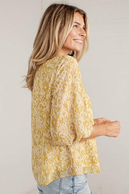 A woman wearing a yellow and white patterned blouse with three-quarter sleeves, shown from a side-back angle against a light gray brick wall.
