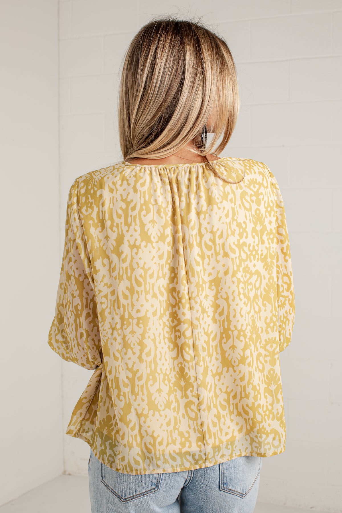 A woman viewed from behind wearing a yellow and white patterned blouse with three-quarter sleeves and light blue jeans, standing against a white paneled wall.