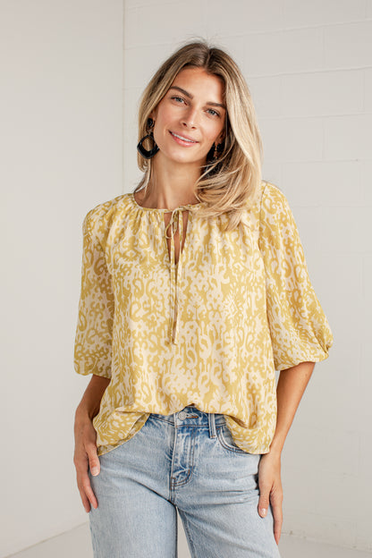 A woman wearing a yellow and white patterned blouse with three-quarter sleeves and a tie neckline, paired with light-wash blue jeans, standing against a white brick wall.
