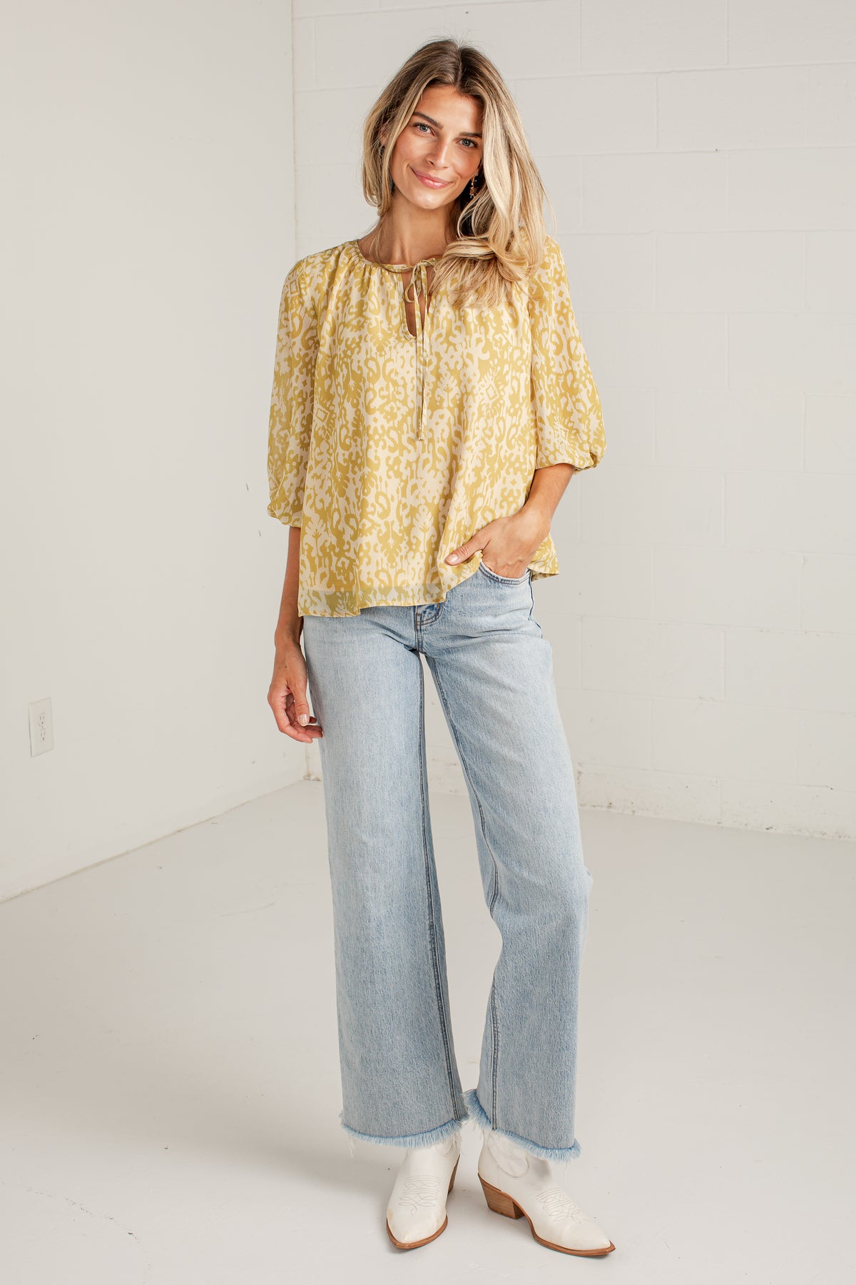 A woman modeling a yellow and white floral patterned blouse with elbow-length puff sleeves, paired with light-wash wide-leg jeans and white ankle boots, standing in a bright white studio.