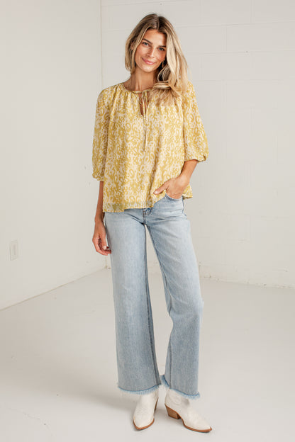 A woman modeling a yellow and white floral patterned blouse with elbow-length puff sleeves, paired with light-wash wide-leg jeans and white ankle boots, standing in a bright white studio.