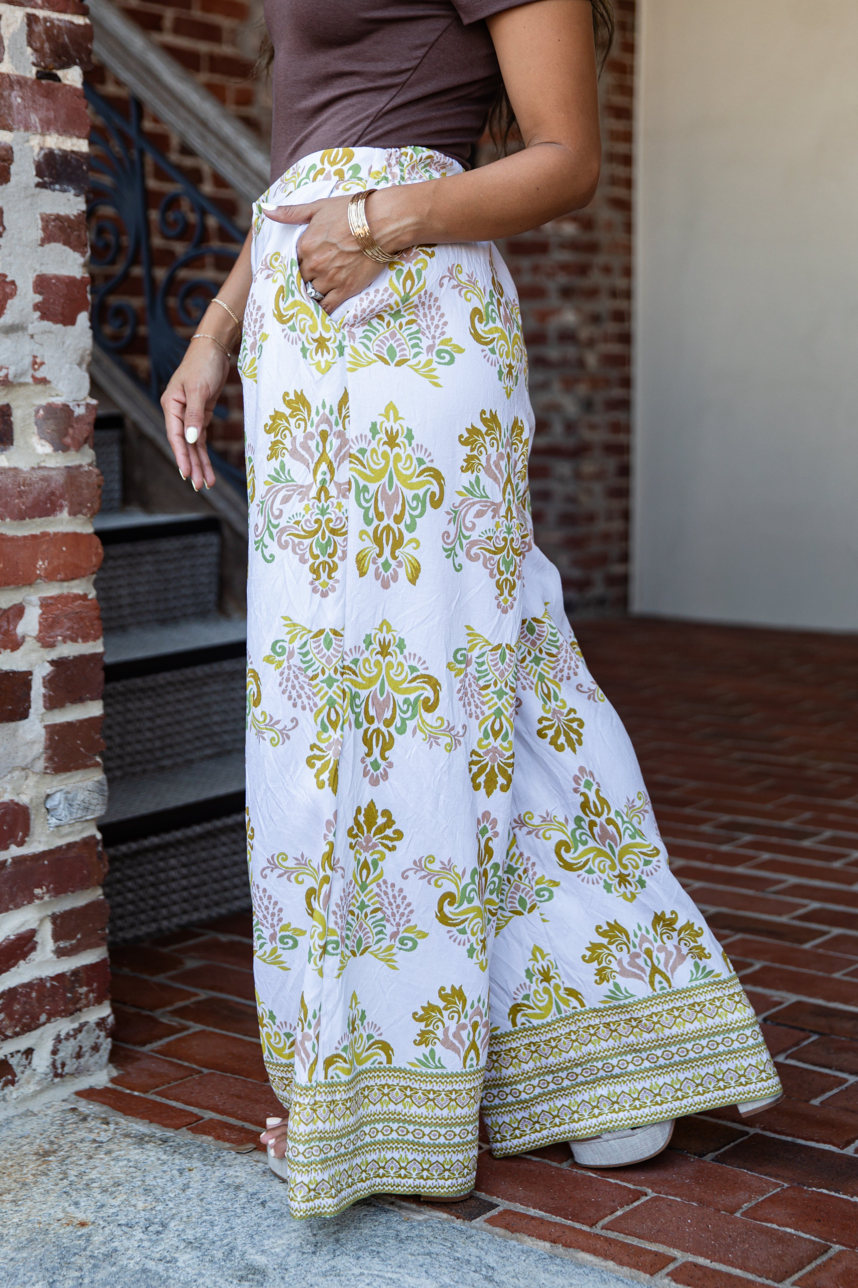 A person wearing a white maxi skirt with a colorful green, yellow, and pink floral paisley pattern and a decorative border, paired with a brown top, standing on a brick patio.