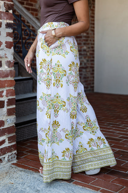 A person wearing a white maxi skirt with a colorful green, yellow, and pink floral paisley pattern and a decorative border, paired with a brown top, standing on a brick patio.