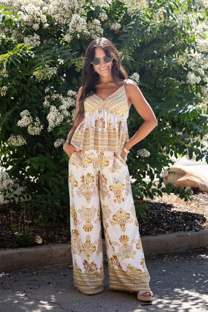 A woman wearing a sleeveless bohemian-style jumpsuit with gold and brown patterns on a white background, standing outdoors in front of a leafy bush with white flowers.