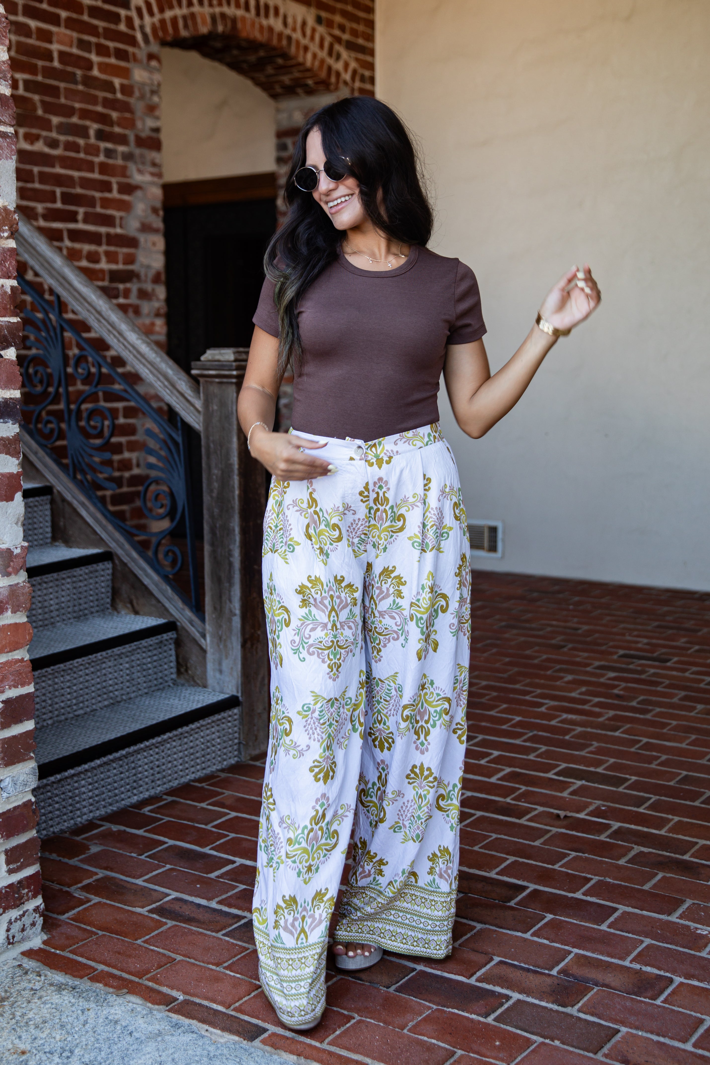 A woman wearing a brown short-sleeve t-shirt and white wide-leg pants with a green and yellow botanical pattern, standing on a brick patio.