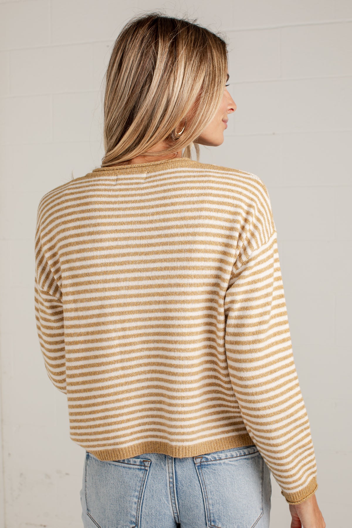 A woman shown from the back wearing a beige and white striped knit sweater with light blue jeans, standing against a white brick wall.