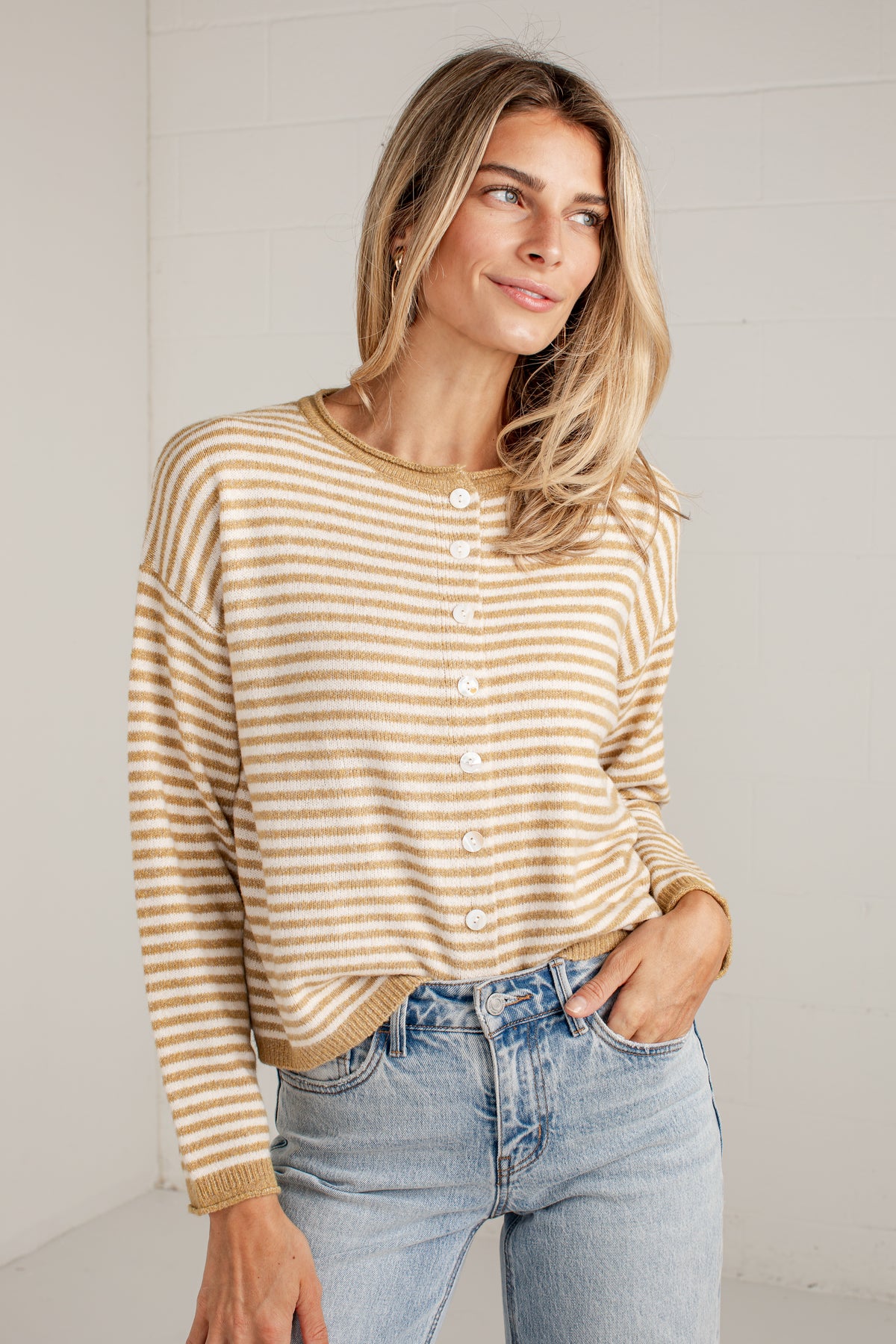 A woman wearing a beige and white striped long-sleeve henley top with white buttons, paired with light-wash blue jeans, standing against a white paneled wall.