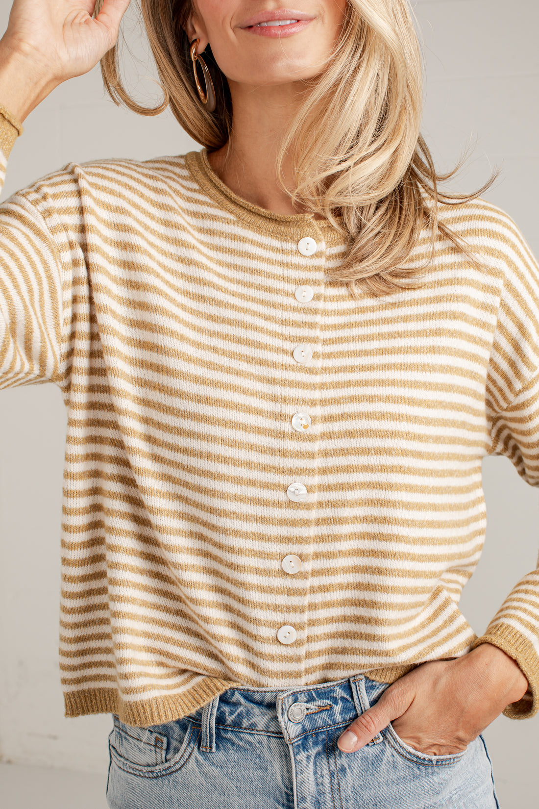 A woman wearing a beige and tan striped button-up cardigan sweater with white buttons, paired with light-wash blue jeans.
