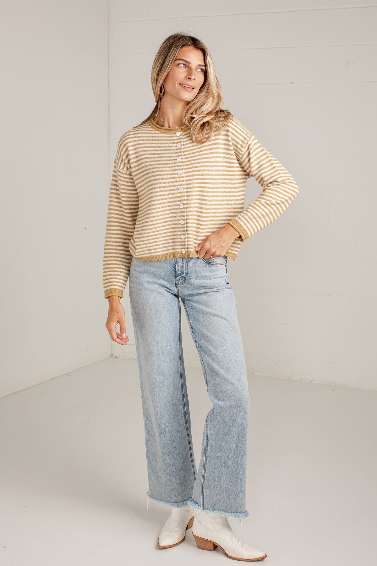 A woman modeling a beige and white striped long-sleeve knit cardigan with button-front detailing, paired with light-wash wide-leg jeans and white ankle boots, standing against a white studio wall.
