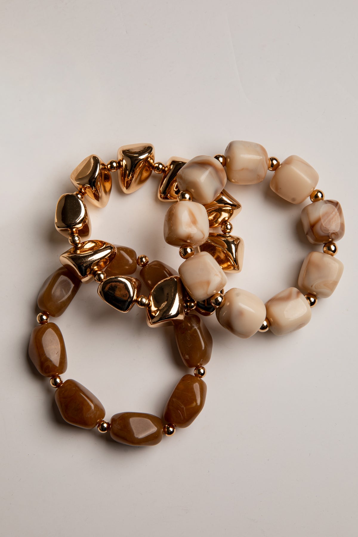 Two beaded stretch bracelets with metallic gold star and heart charms, one featuring smooth cream beads and the other featuring marbled beige and brown beads, displayed on a light gray background.