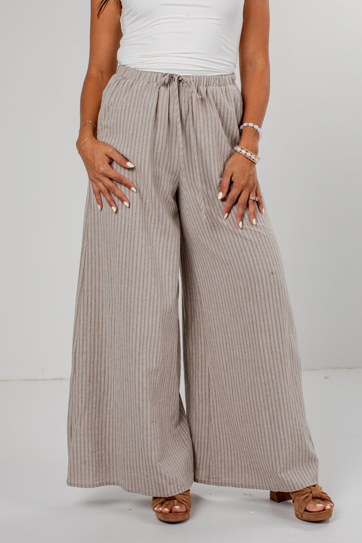 A person wearing wide-leg pleated pants in a light taupe-beige color, styled with a white top and brown sandals against a plain light gray studio background.