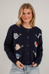 A woman wearing a navy blue knit cardigan with colorful fish and sea-creature motifs, paired with light-wash jeans, standing against a white brick wall.
