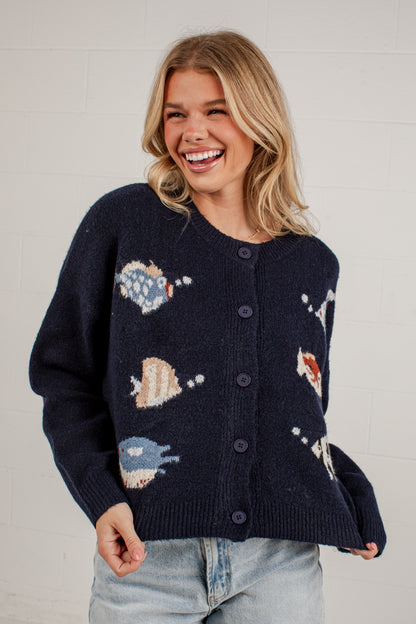 A woman smiling while wearing a navy blue button-up cardigan with knitted fish motifs in blue, beige, and red, paired with light-wash jeans against a white brick wall.