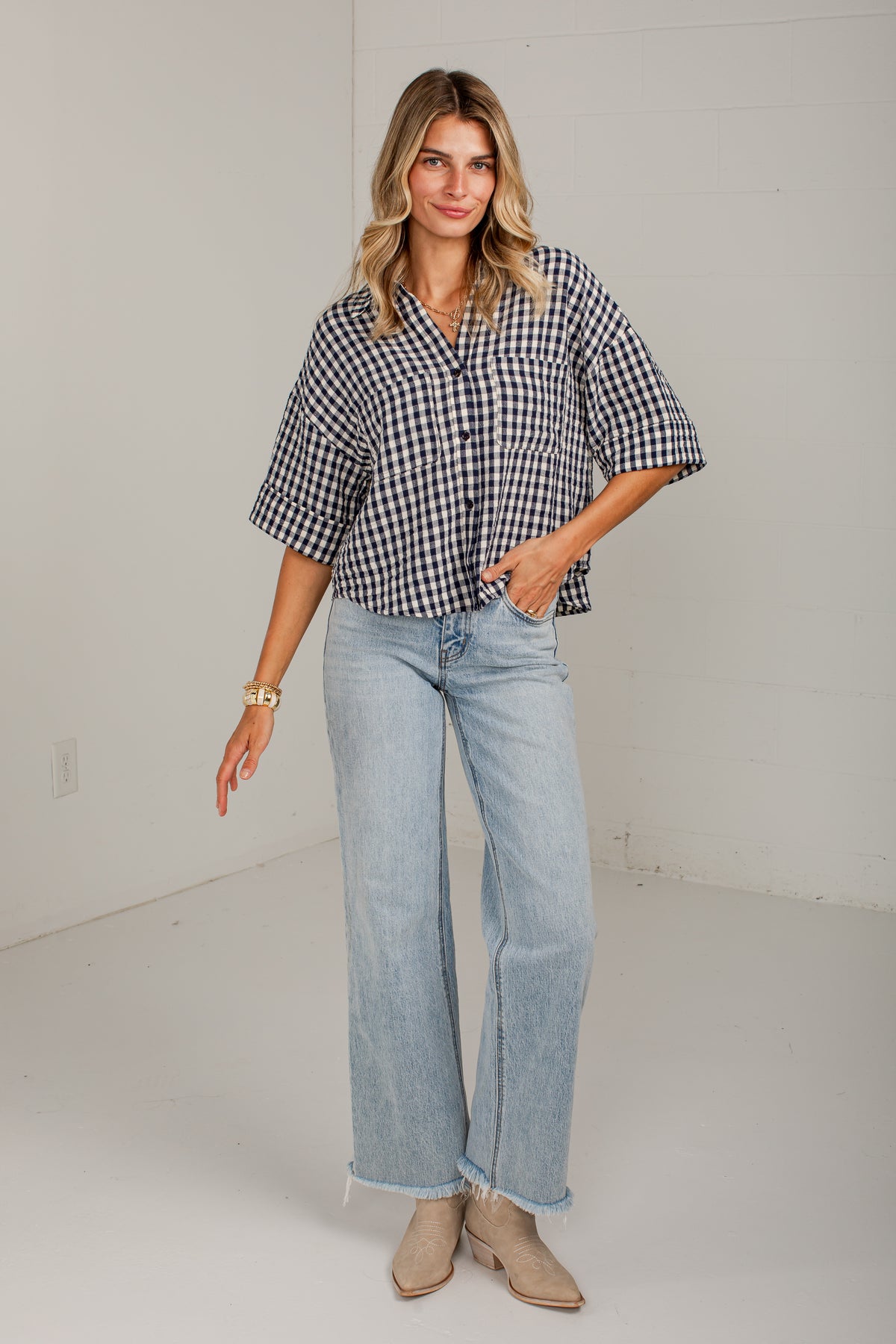 A woman modeling a black and white gingham short-sleeve button-up shirt with light-wash wide-leg jeans and beige ankle boots, standing in a bright white studio.