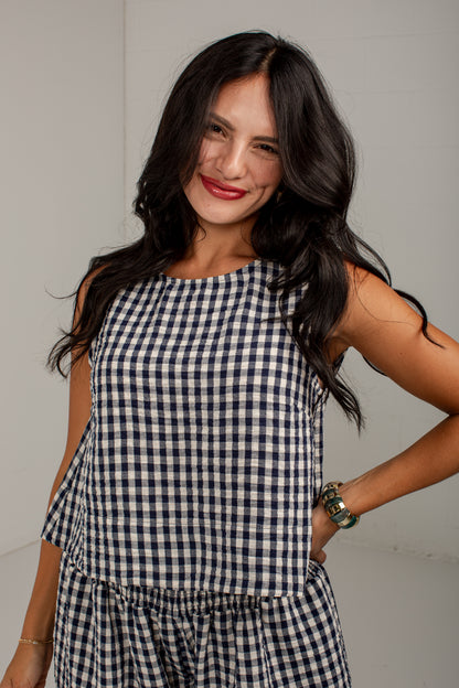A woman with long dark hair wearing a sleeveless navy and white gingham check top, posing against a plain light gray wall.