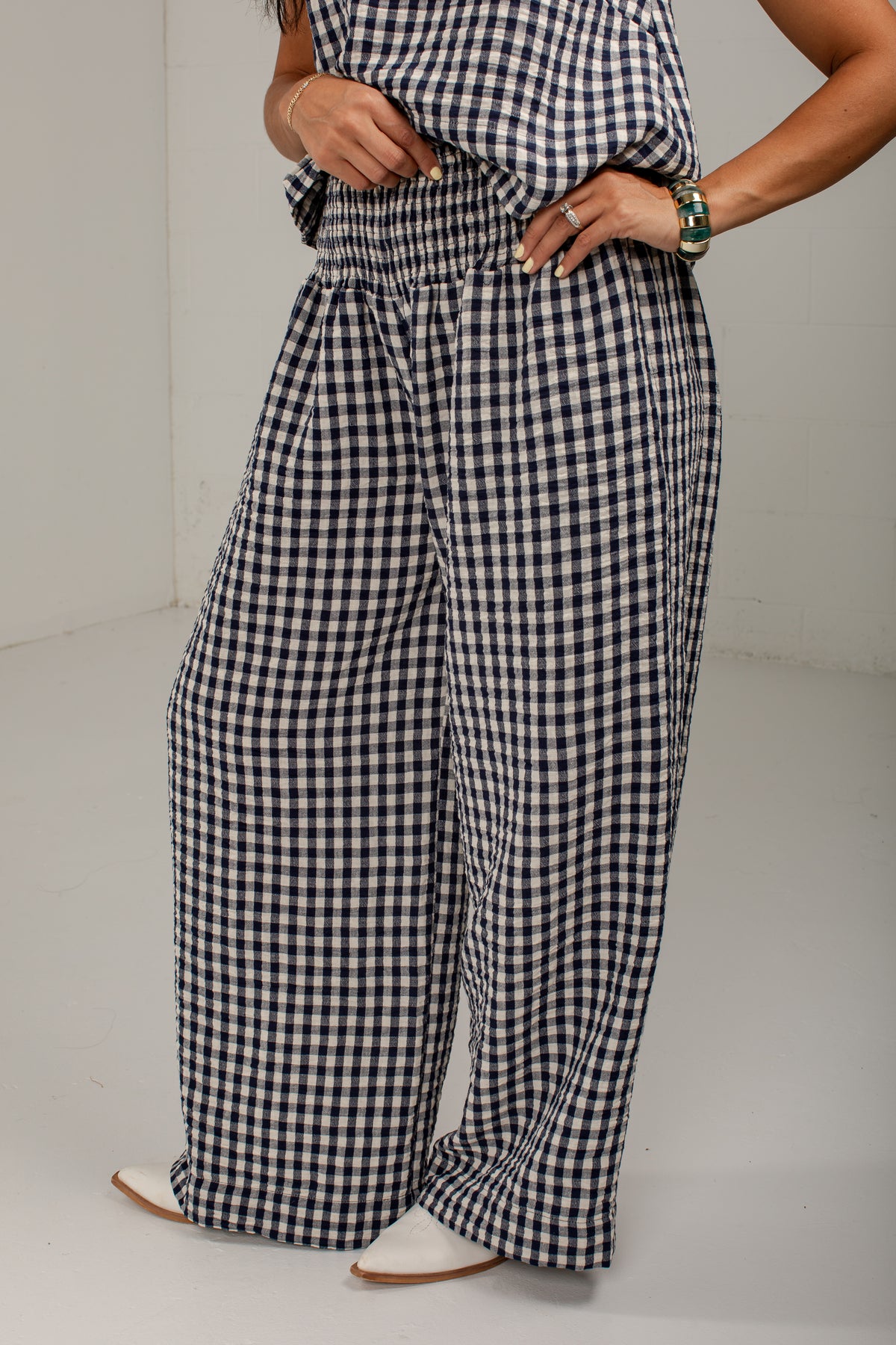 A person wearing loose, wide-leg gingham pants in a black and white check pattern, paired with a matching gingham top and white pointed shoes, photographed in a plain indoor setting.