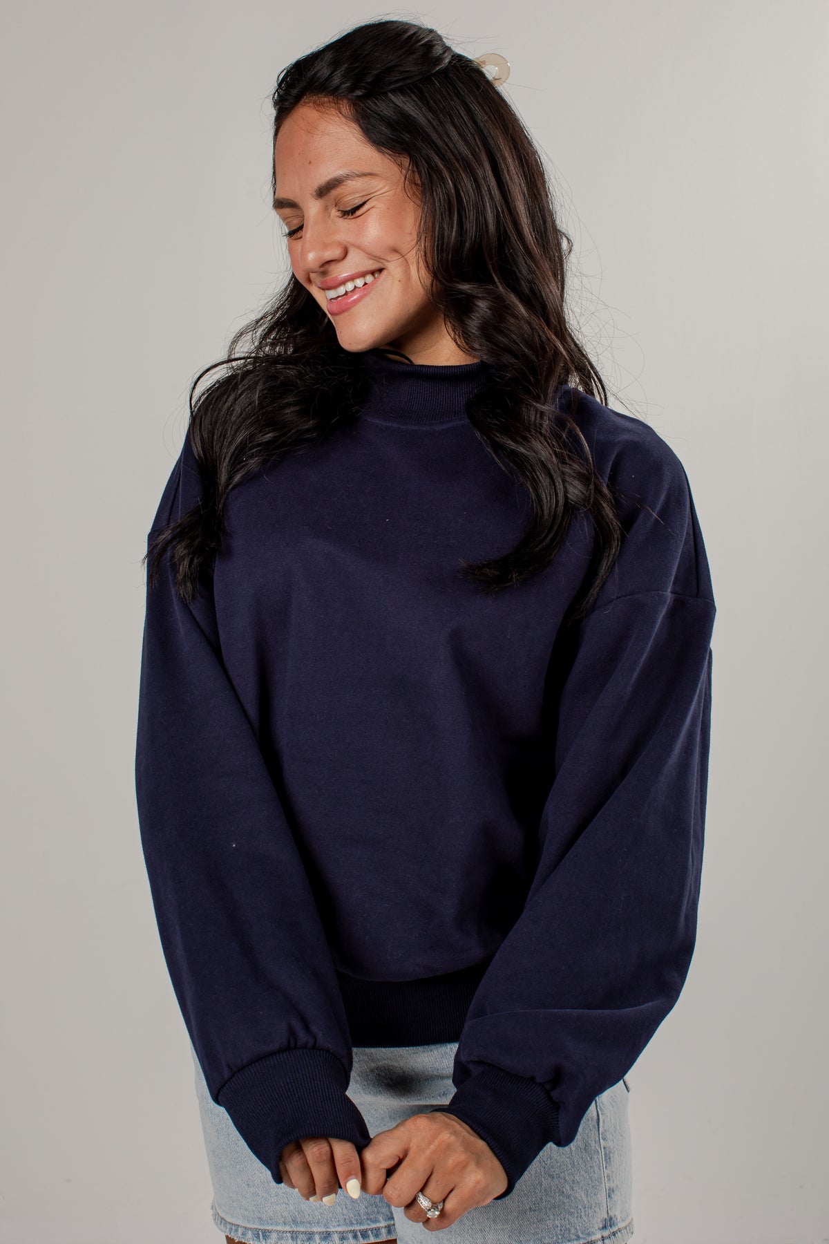 A woman wearing a dark navy blue mockneck sweatshirt and light wash denim jeans, standing against a neutral light gray studio background.