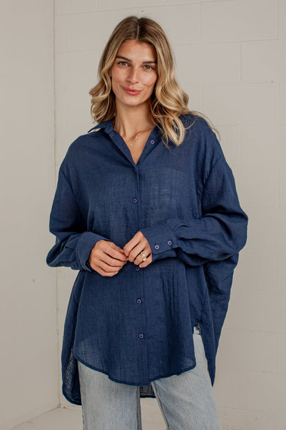 A woman wearing a dark blue, oversized long-sleeve linen button-up shirt with light-wash jeans, standing against a white brick wall.