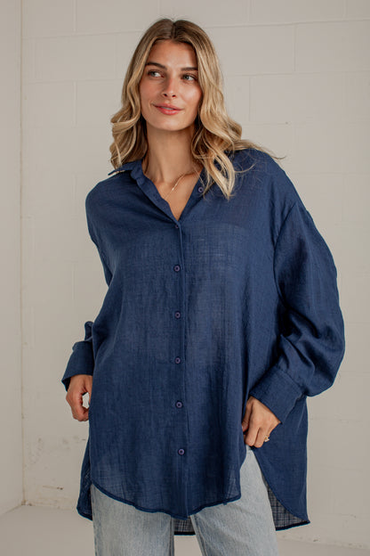 A woman wearing an oversized dark blue linen button-up shirt with light-wash jeans, standing against a white brick wall.
