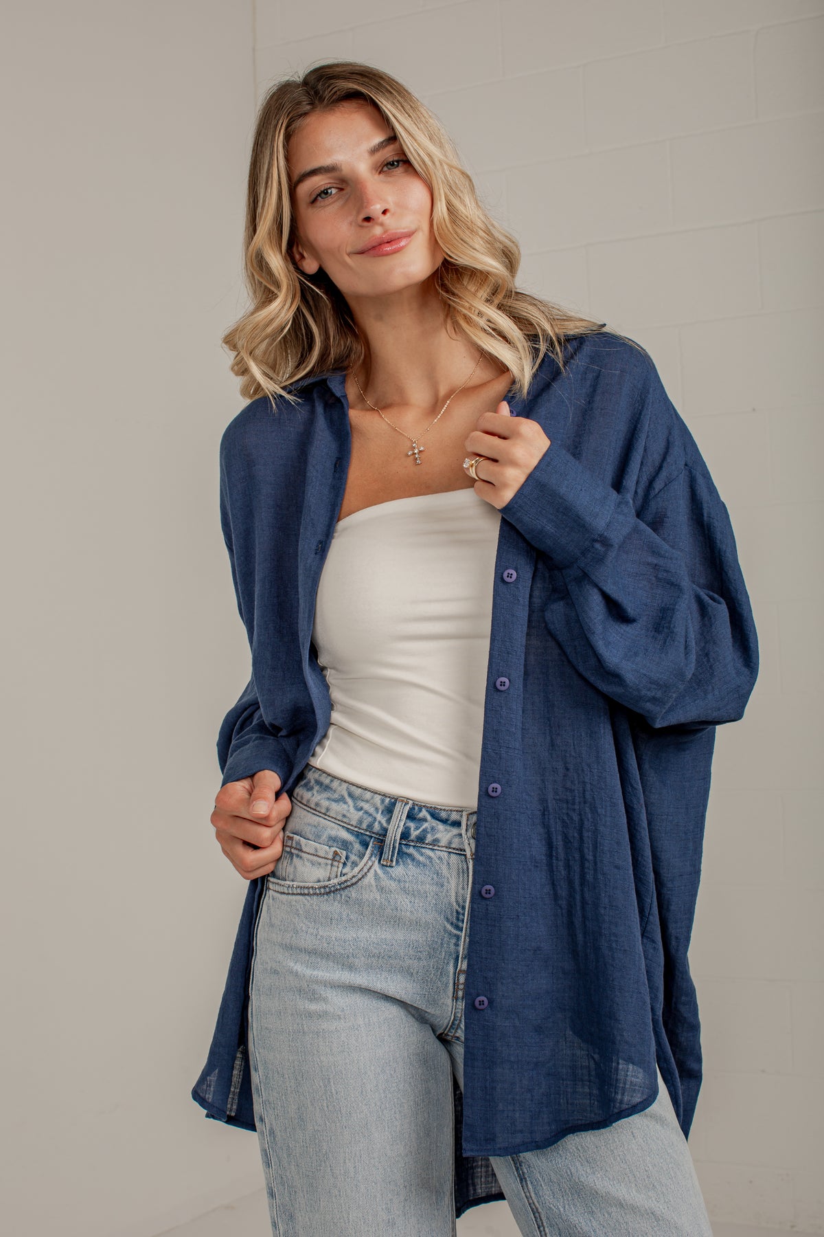 A woman wearing a loose, oversized blue button-up shirt over a white tube top and light-wash blue jeans, standing against a light gray wall.