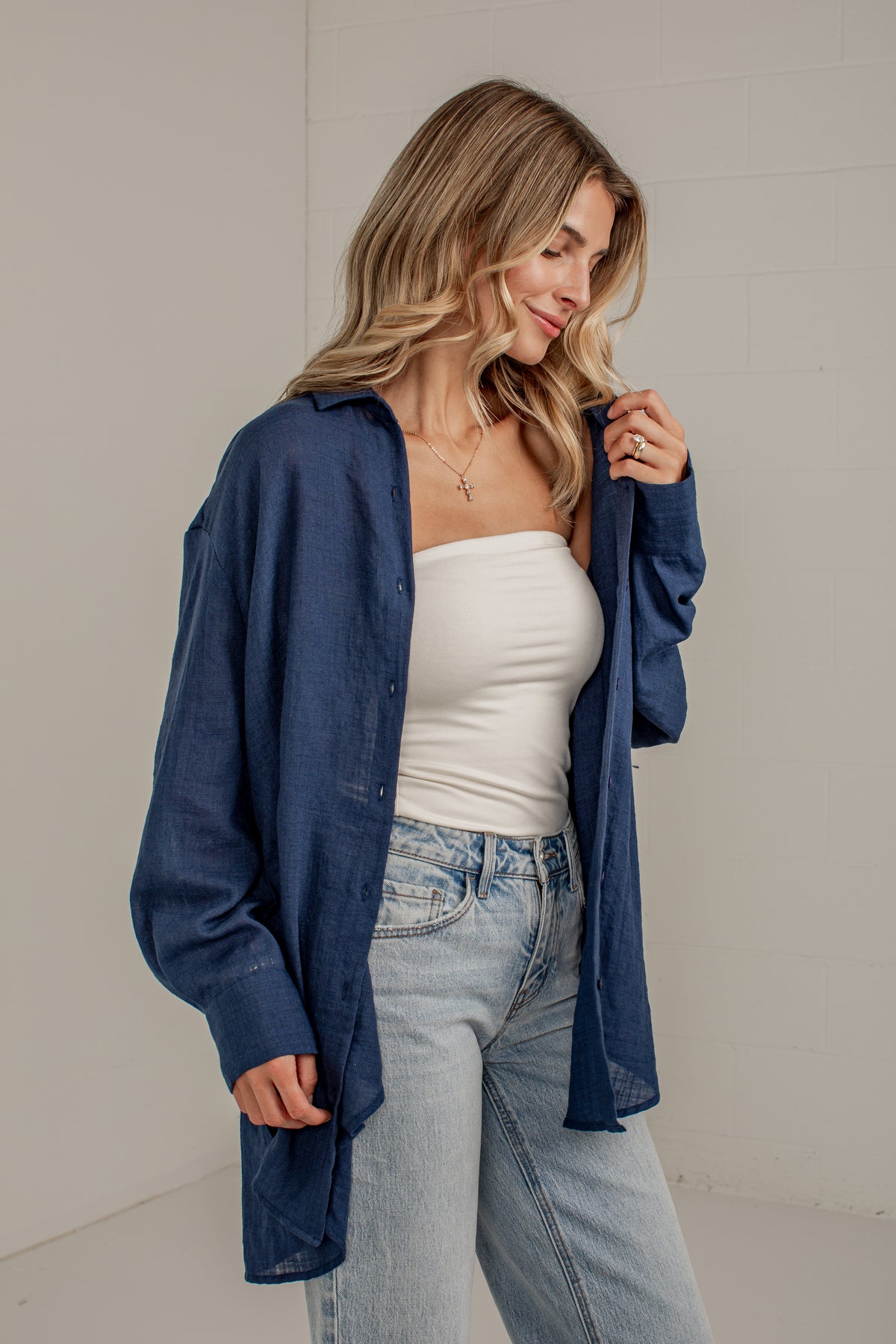 A woman wearing a navy blue button-up shirt over a white strapless top and light-wash blue jeans, standing against a light gray wall.