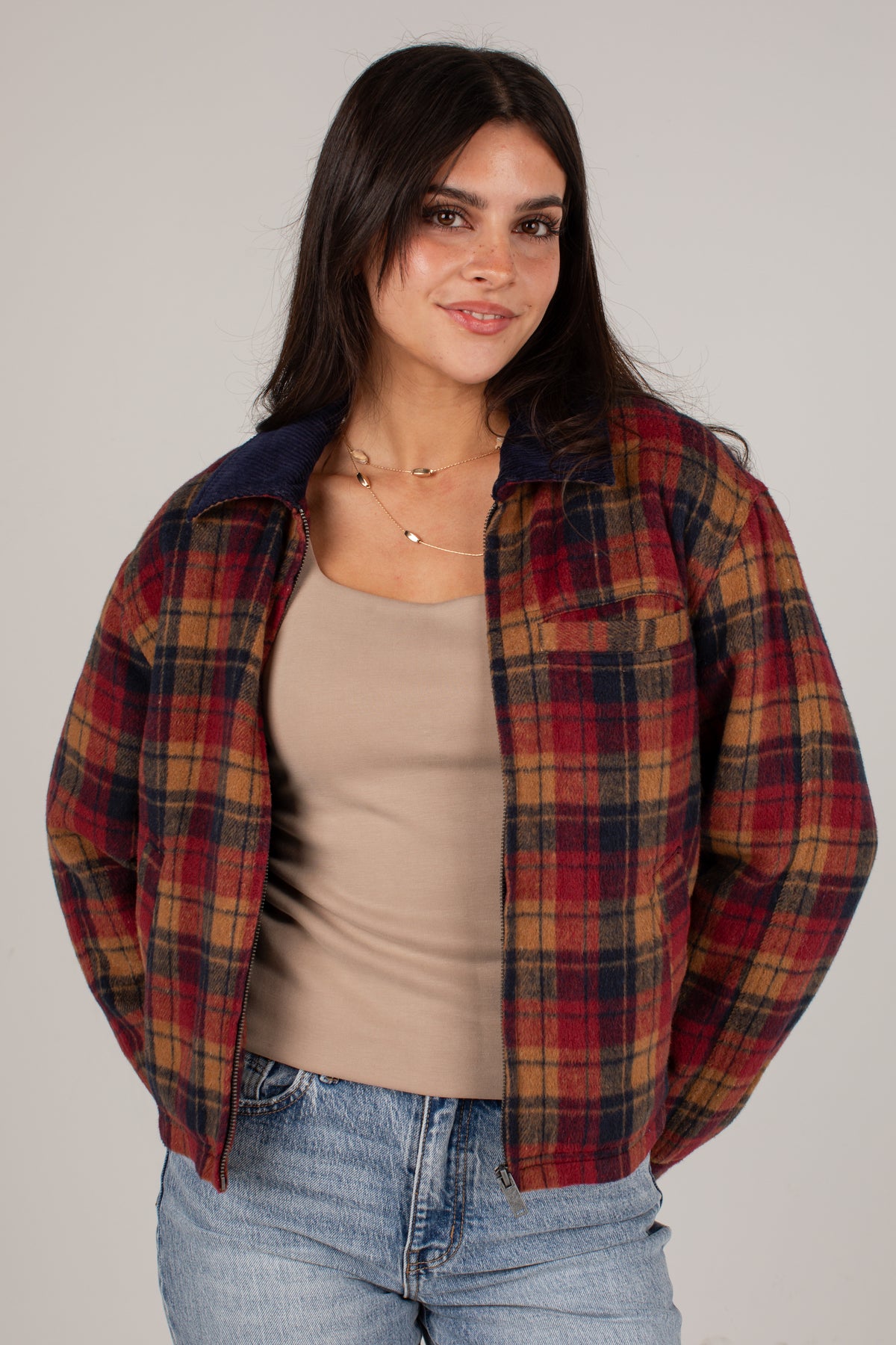 A woman wearing a red, brown, and navy plaid overshirt over a beige tank top and blue jeans, photographed against a plain light gray studio background.