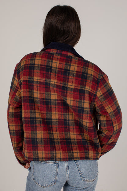 A person viewed from the back wearing a red, navy, and tan plaid flannel-style jacket with a dark navy collar, paired with light blue jeans, against a plain light gray studio background.