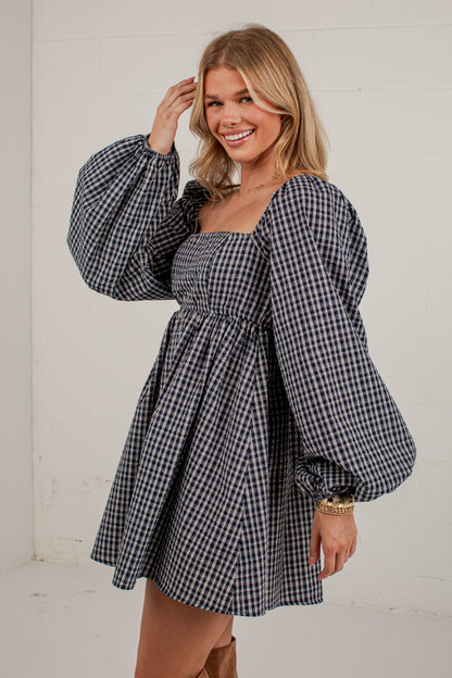 A woman modeling a black and white gingham mini dress with voluminous balloon sleeves, a square neckline, and a gathered empire waist, set against a white brick wall.