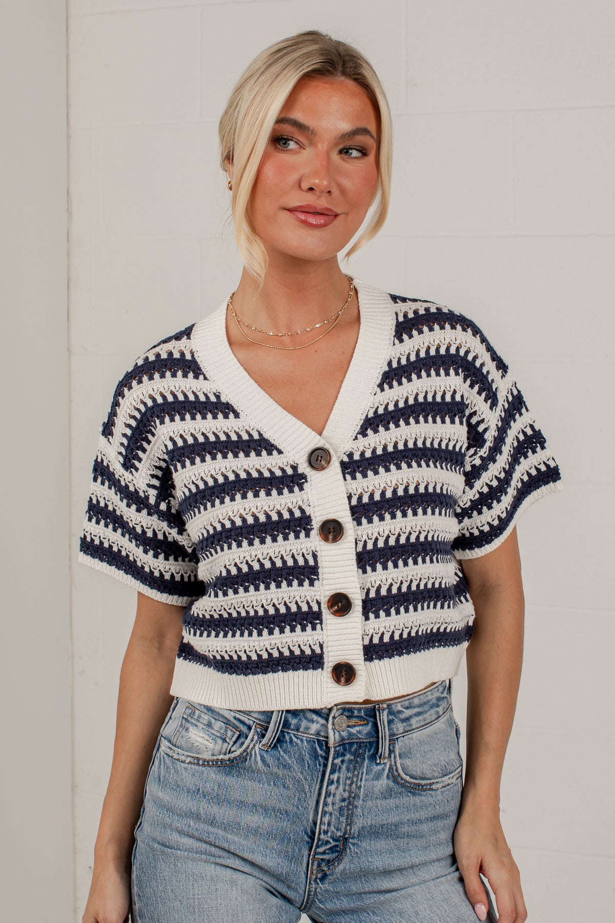 A woman wearing a short-sleeve knit cardigan with navy and cream horizontal stripes and brown buttons, paired with light wash blue jeans, standing against a white brick wall.