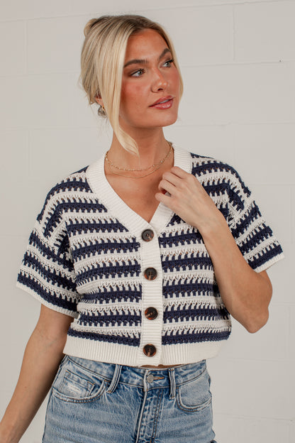 A woman wearing a short-sleeve knit cardigan with navy and cream horizontal stripes, brown buttons, and a V-neck, paired with blue jeans against a white brick wall.
